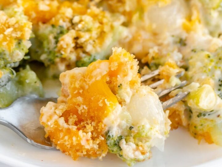 Close-up of a fork lifting a portion of a cheesy broccoli and cauliflower casserole. The dish is topped with breadcrumbs, visible through melted cheese, with vibrant green broccoli and orange mixed in a creamy sauce on a white plate.
