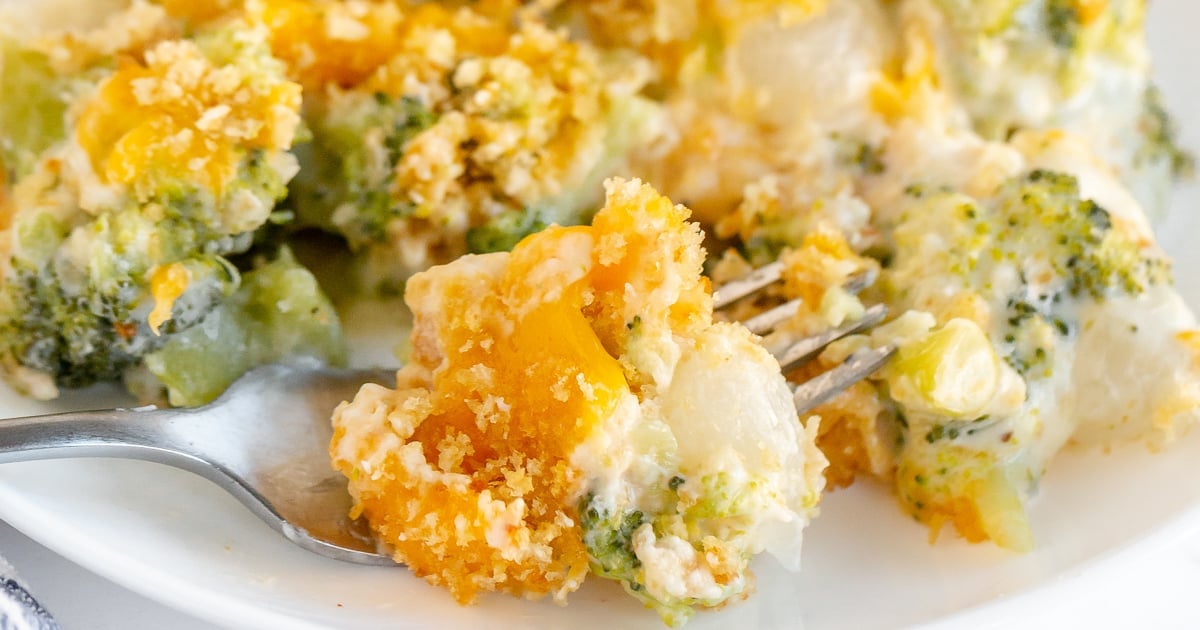 Close-up of a fork lifting a portion of a cheesy broccoli and cauliflower casserole. The dish is topped with breadcrumbs, visible through melted cheese, with vibrant green broccoli and orange mixed in a creamy sauce on a white plate.