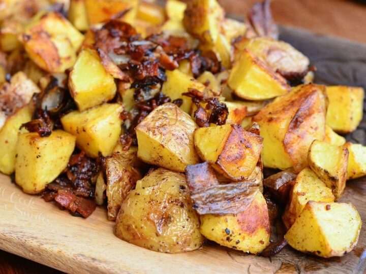 A wooden tray filled with roasted yellow potatoes, some with crispy, browned skin, and bits of caramelized onions. The texture appears crispy and seasoned, displaying a golden-brown hue.