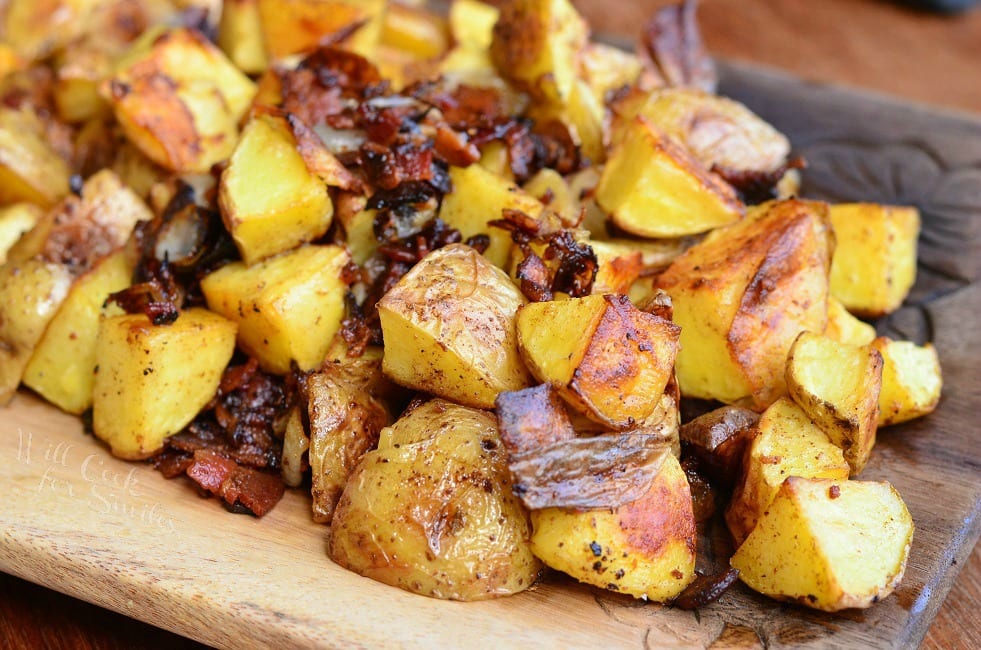 A wooden tray filled with roasted yellow potatoes, some with crispy, browned skin, and bits of caramelized onions. The texture appears crispy and seasoned, displaying a golden-brown hue.