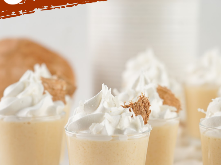 Five small dessert cups filled with creamy coconut jello shots, each topped with whipped cream and a sprinkle of brown crumb. The background features a coconut and stacks of paper cups. The text at the top reads Coconut Jello Shots.