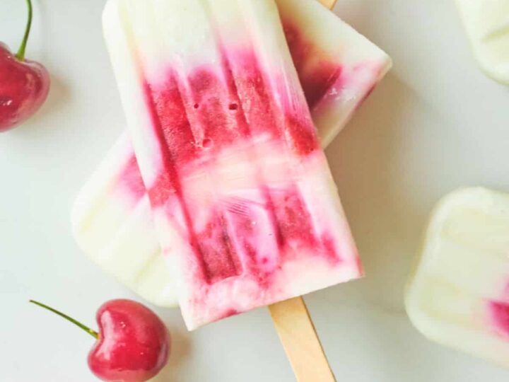 Three cherry coconut milk popsicles on a white surface, surrounded by fresh cherries. The popsicles have a swirled pink and white appearance. Text at the top reads Cherry Coconut Milk Popsicles and EverydaySouthwest.com at the bottom.