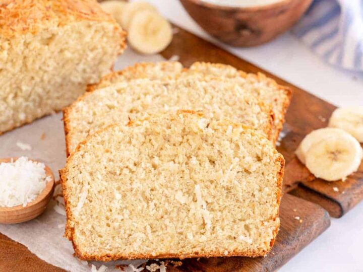 Slices of banana bread with visible shredded coconut on a wooden board. Whole loaf and banana slices are in the background, alongside a small bowl of shredded coconut.