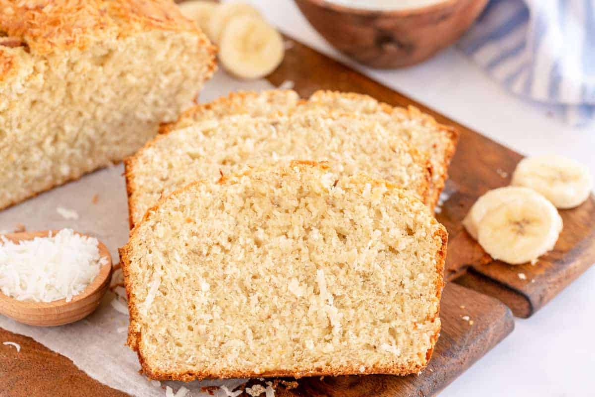 Slices of banana bread with visible shredded coconut on a wooden board. Whole loaf and banana slices are in the background, alongside a small bowl of shredded coconut.