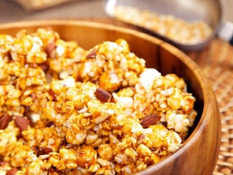 A wooden bowl filled with caramel popcorn mixed with nuts is placed on a woven placemat. In the background, theres a scoop and an open paper bag filled with popcorn kernels.