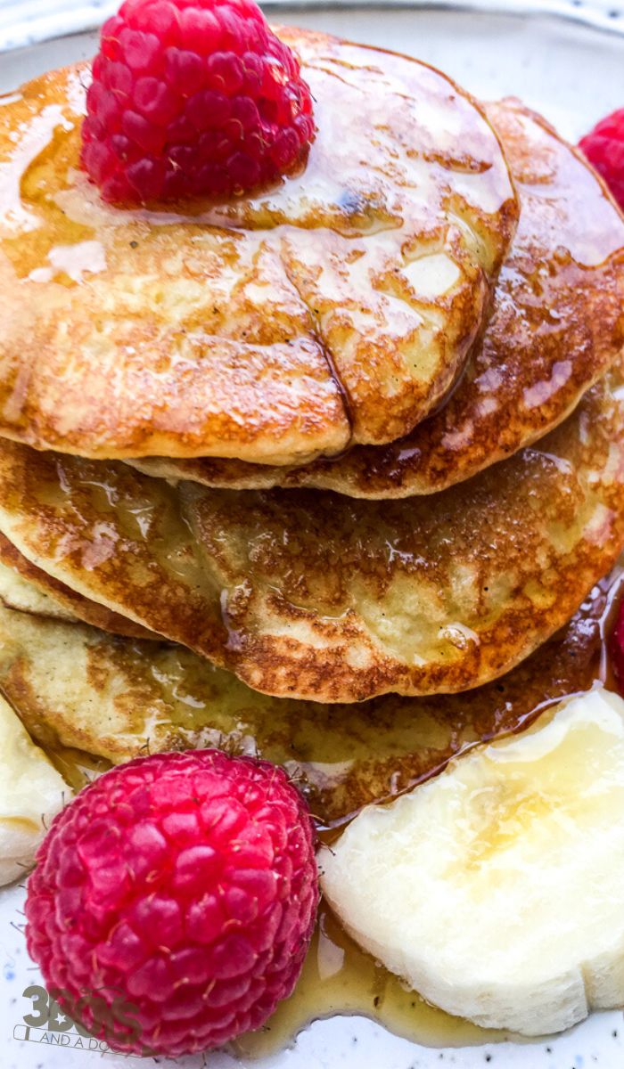 A stack of golden brown coconut flour pancakes topped with fresh raspberries, sliced bananas, and drizzled with syrup, served on a plate.