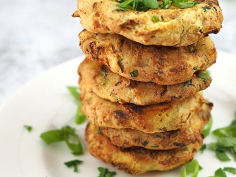 A stack of golden-brown colcannon cakes topped with fresh green herbs, sitting on a white plate. Additional herbs are scattered around the plate. Text in the image reads vegan + gluten-free Colcannon Cakes, Diannes Vegan Kitchen.