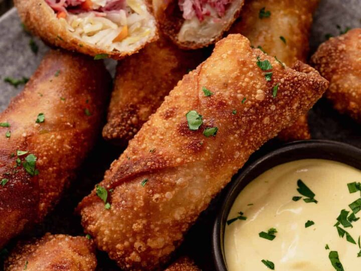 A plate of crispy egg rolls filled with meat and vegetables, garnished with chopped herbs. A small bowl of creamy dipping sauce is beside the egg rolls on the plate.