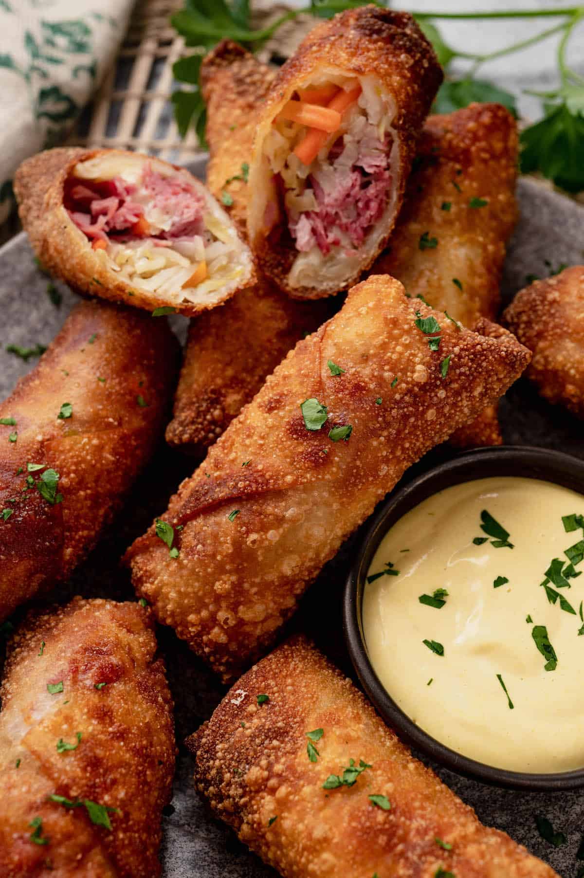 A plate of crispy egg rolls filled with meat and vegetables, garnished with chopped herbs. A small bowl of creamy dipping sauce is beside the egg rolls on the plate.