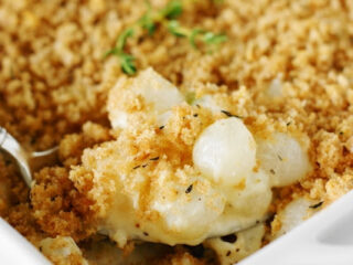 Close-up of a white casserole dish filled with a creamy mixture topped with golden-brown breadcrumbs. The dish is garnished with a sprig of fresh thyme, and a spoon is partially scooping out the contents.