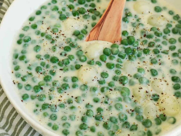 A bowl filled with creamy peas and pearl onions in a white sauce, garnished with cracked black pepper. A wooden spoon rests in the bowl, and a striped green and white cloth is visible beside it.