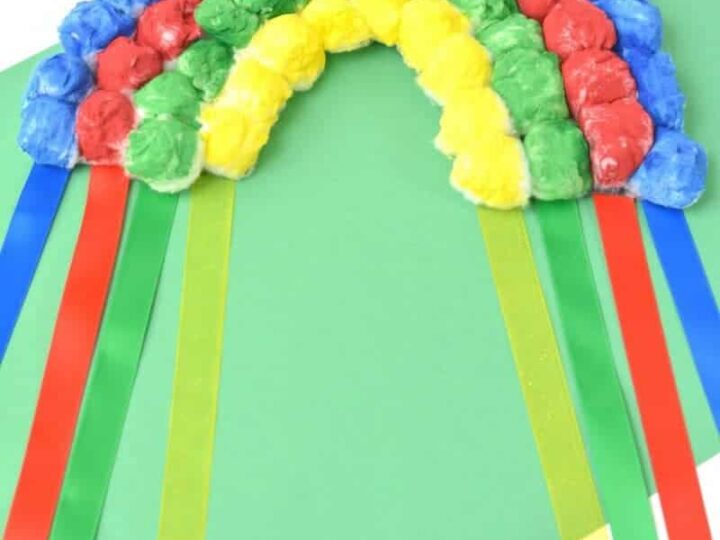 A craft rainbow made from colored tissue paper balls, arranged in arcs and glued onto a green background. Four ribbons, in red, blue, yellow, and green, hang down from the rainbow. A small circle logo at the bottom reads The Inspiration Edit.