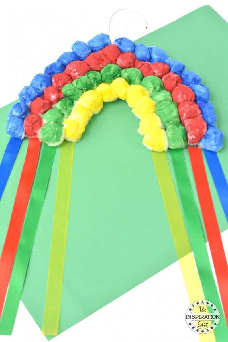 A craft rainbow made from colored tissue paper balls, arranged in arcs and glued onto a green background. Four ribbons, in red, blue, yellow, and green, hang down from the rainbow. A small circle logo at the bottom reads The Inspiration Edit.