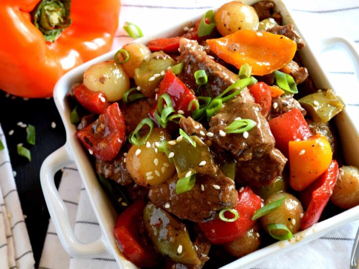 A dish of beef stir fry served in a square white dish. The stir fry includes bell peppers, pearl onions, and green onions, topped with sesame seeds. A sliced orange bell pepper is visible near the dish.