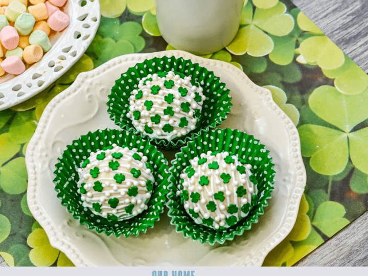 Three white chocolate-covered treats with green clover sprinkles sit in green polka dot cupcake liners on a decorative plate. Nearby, there is a cup and a plate of pastel-colored candies. The setting includes a clover-themed placemat.