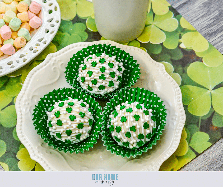 Three white chocolate-covered treats with green clover sprinkles sit in green polka dot cupcake liners on a decorative plate. Nearby, there is a cup and a plate of pastel-colored candies. The setting includes a clover-themed placemat.