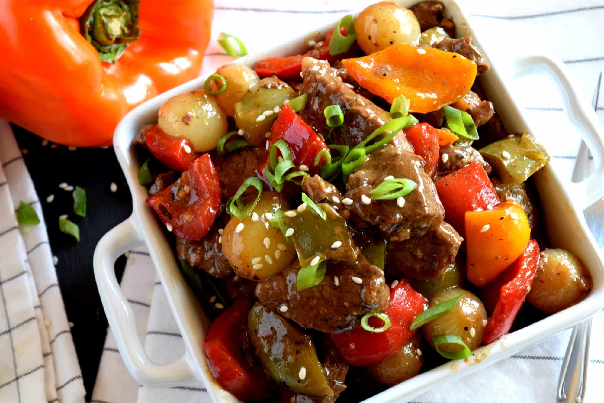 A dish of beef stir fry served in a square white dish. The stir fry includes bell peppers, pearl onions, and green onions, topped with sesame seeds. A sliced orange bell pepper is visible near the dish.