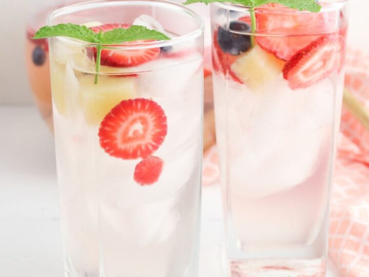 Two glasses of infused water with ice cubes, sliced strawberries, blueberries, and mint leaves. The glasses are on a white surface near a pink cloth. A pitcher of the same beverage is partially visible in the background.