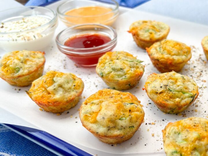 Mini muffins with herbs and melted cheese on a white tray, accompanied by bowls of dipping sauces: creamy white, orange, and red. The tray is placed on a blue and white checkered cloth.