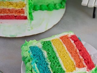A slice of rainbow cake on a white plate, showing five colorful layers: blue, green, yellow, orange, and red. The cake is frosted with green icing and has decorative green piping along the edges. A partially sliced whole cake is in the background.