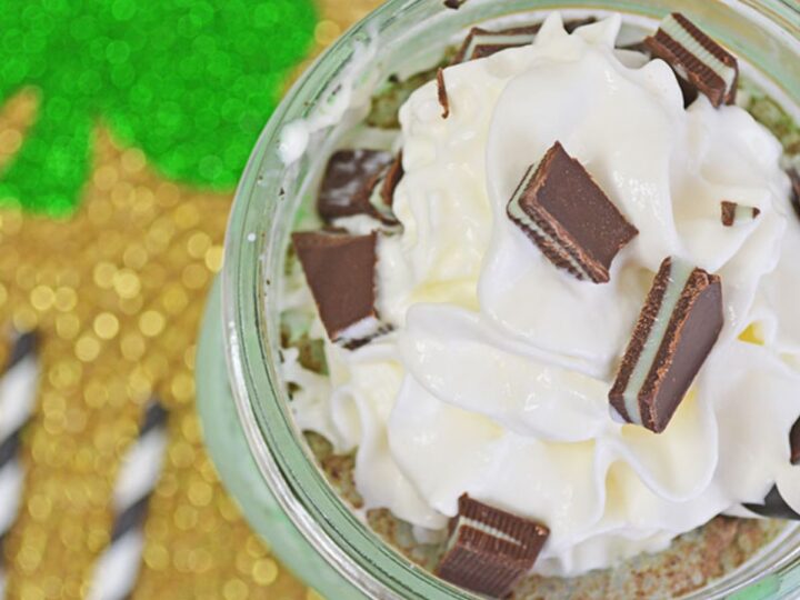 Whipped cream topped with chocolate pieces in a jar, sitting on a gold and green surface with a clover design. Two striped straws are visible beside the jar.