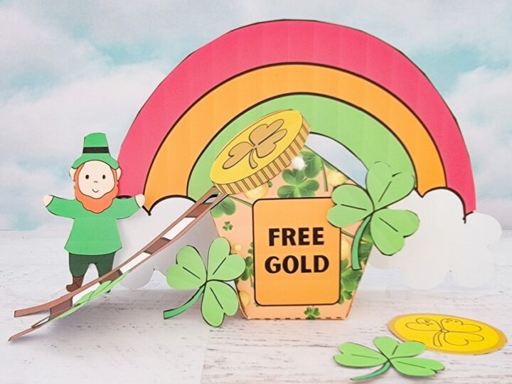 A paper craft scene with a leprechaun standing on a rainbow leading to a jar labeled Free Gold. Shamrocks are scattered around, and white clouds frame the rainbow against a cloudy sky backdrop.