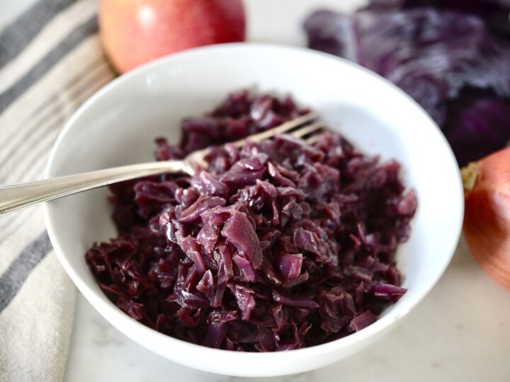 A white bowl filled with cooked red cabbage sits invitingly, with a fork resting inside. In the background, a striped kitchen towel complements a whole red apple, part of a red cabbage, and an unpeeled red onion on a marble surface—perfect elements for recipes that use cabbage.
