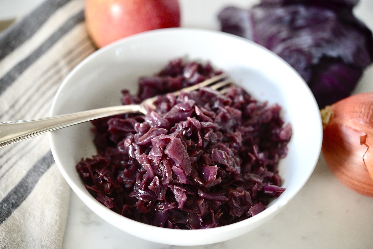 A white bowl filled with cooked red cabbage sits invitingly, with a fork resting inside. In the background, a striped kitchen towel complements a whole red apple, part of a red cabbage, and an unpeeled red onion on a marble surface—perfect elements for recipes that use cabbage.