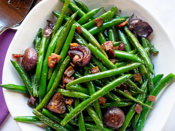 A white bowl filled with fresh green beans, small roasted onions, and crispy bacon pieces, all glistening with seasoning. Next to the bowl are a serving spoon and fork on a purple napkin.