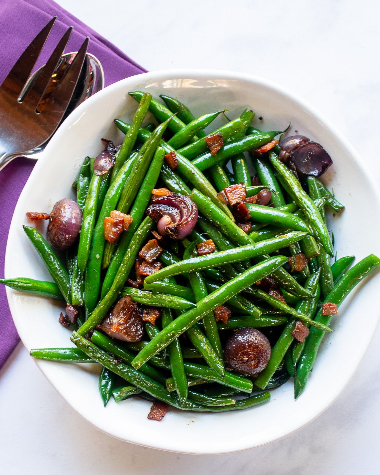 A white bowl filled with fresh green beans, small roasted onions, and crispy bacon pieces, all glistening with seasoning. Next to the bowl are a serving spoon and fork on a purple napkin.
