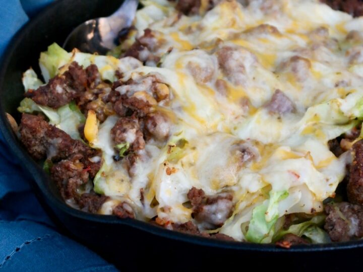 Cheesy beef and cabbage dish in a black skillet, featuring ground beef, shredded cabbage, and melted cheese on top. A spoon is placed in the skillet, and a blue cloth is visible underneath.
