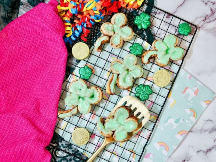 A wire rack holds shamrock-shaped cookies with green icing and green shamrock candies. A pink cloth and colorful streamers are nearby. A spatula rests on the rack, and a unicorn-themed paper is in the background on a marble surface.