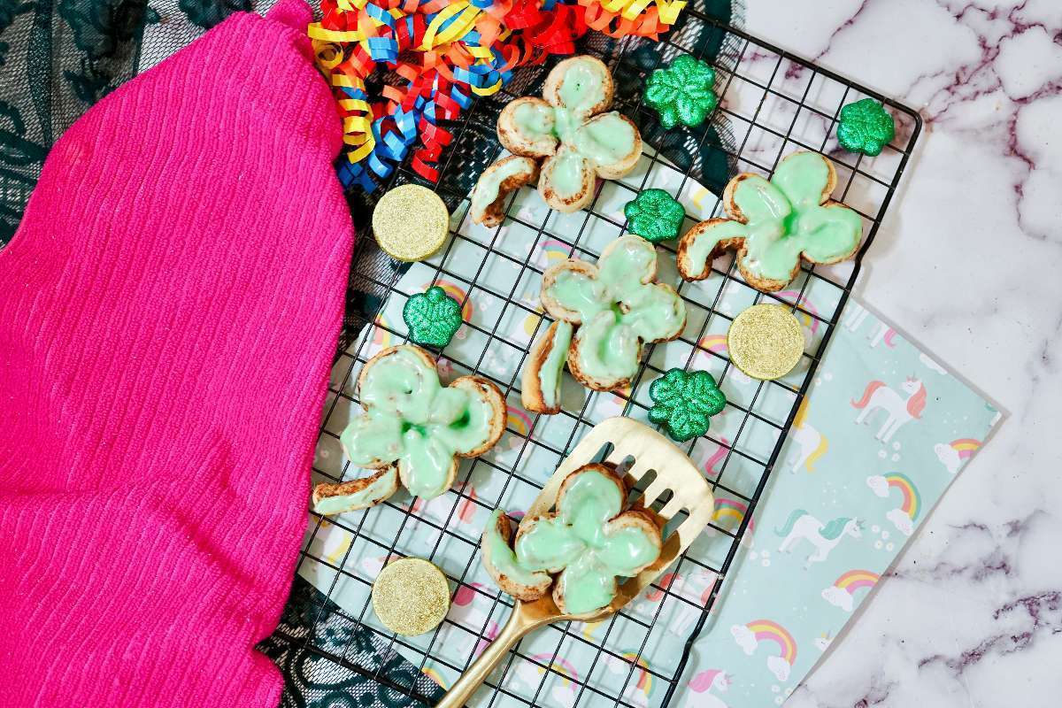 A wire rack holds shamrock-shaped cookies with green icing and green shamrock candies. A pink cloth and colorful streamers are nearby. A spatula rests on the rack, and a unicorn-themed paper is in the background on a marble surface.