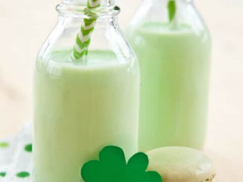 Two glass bottles filled with pale green shamrock shakes, each with a striped straw, are placed on a polka dot napkin. A green shamrock decoration and a macaron accompany them. Text at the top reads Healthy Shamrock Shake.