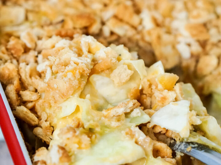 A close-up of a casserole dish filled with creamy, baked cabbage casserole topped with a golden, crunchy crust. A serving spoon is lifting a portion, revealing the creamy layers beneath the crust. The dish sits on a light-colored surface.