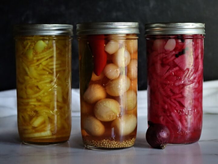 Three jars of pickled vegetables sit on a marble surface against a dark background. From left to right: yellow beans, onions with red peppers, and shredded red cabbage. A small red onion rests in front of the cabbage jar.
