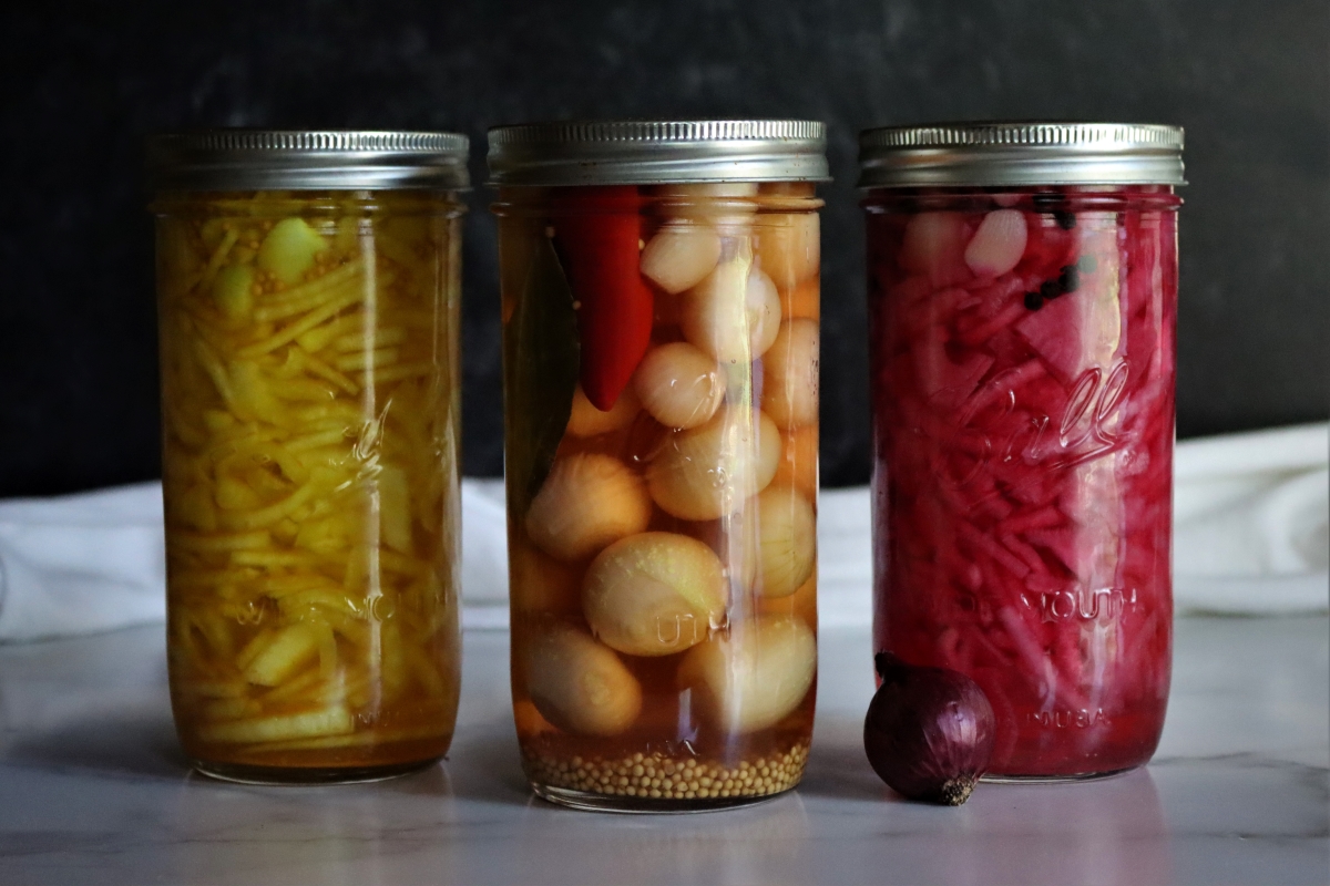 Three jars of pickled vegetables sit on a marble surface against a dark background. From left to right: yellow beans, onions with red peppers, and shredded red cabbage. A small red onion rests in front of the cabbage jar.