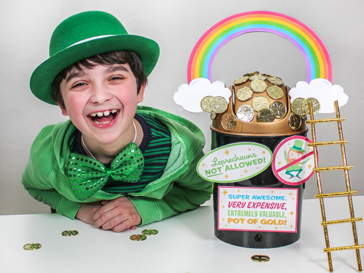 A smiling boy wearing a green hat, jacket, and bowtie sits next to a decorative pot labeled Leprechauns Not Allowed! filled with chocolate coins. A rainbow and ladder add to the festive St. Patricks Day theme.