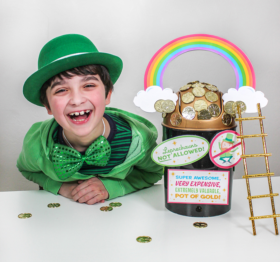 A smiling boy wearing a green hat, jacket, and bowtie sits next to a decorative pot labeled Leprechauns Not Allowed! filled with chocolate coins. A rainbow and ladder add to the festive St. Patricks Day theme.