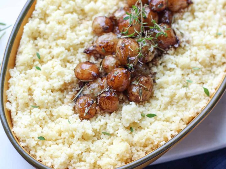 Oval dish filled with couscous topped with roasted onions and garnished with fresh herbs. The dish is placed on a table with a blue napkin and a gold-handled spoon beside it.