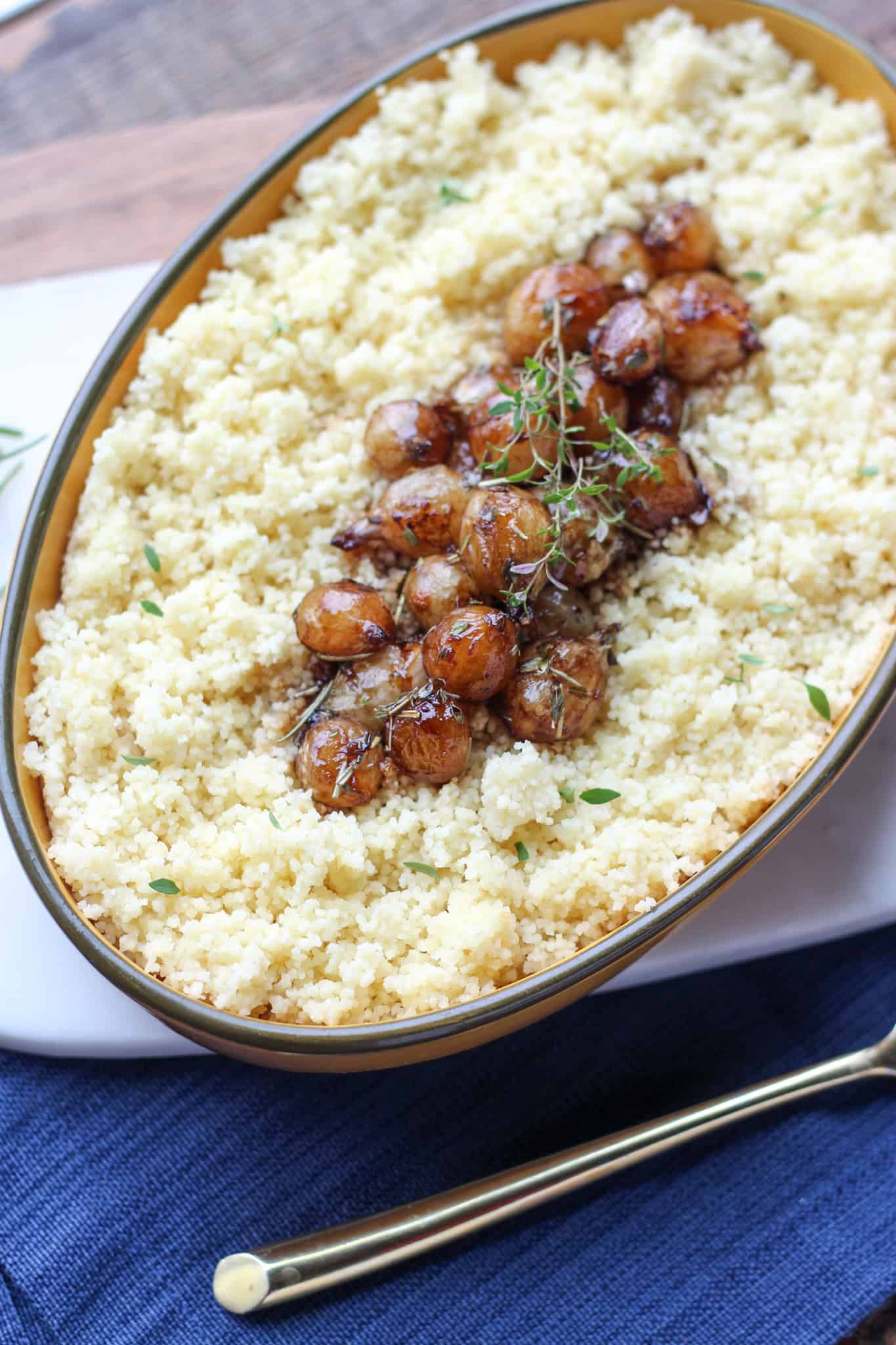 Oval dish filled with couscous topped with roasted onions and garnished with fresh herbs. The dish is placed on a table with a blue napkin and a gold-handled spoon beside it.