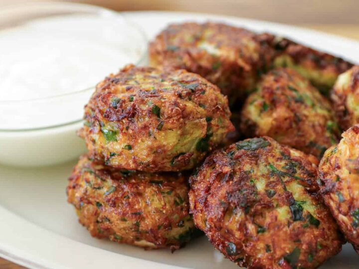 A plate of golden-brown zucchini fritters is stacked next to a small glass bowl filled with white yogurt sauce. The fritters are speckled with green herbs and have a crispy texture.