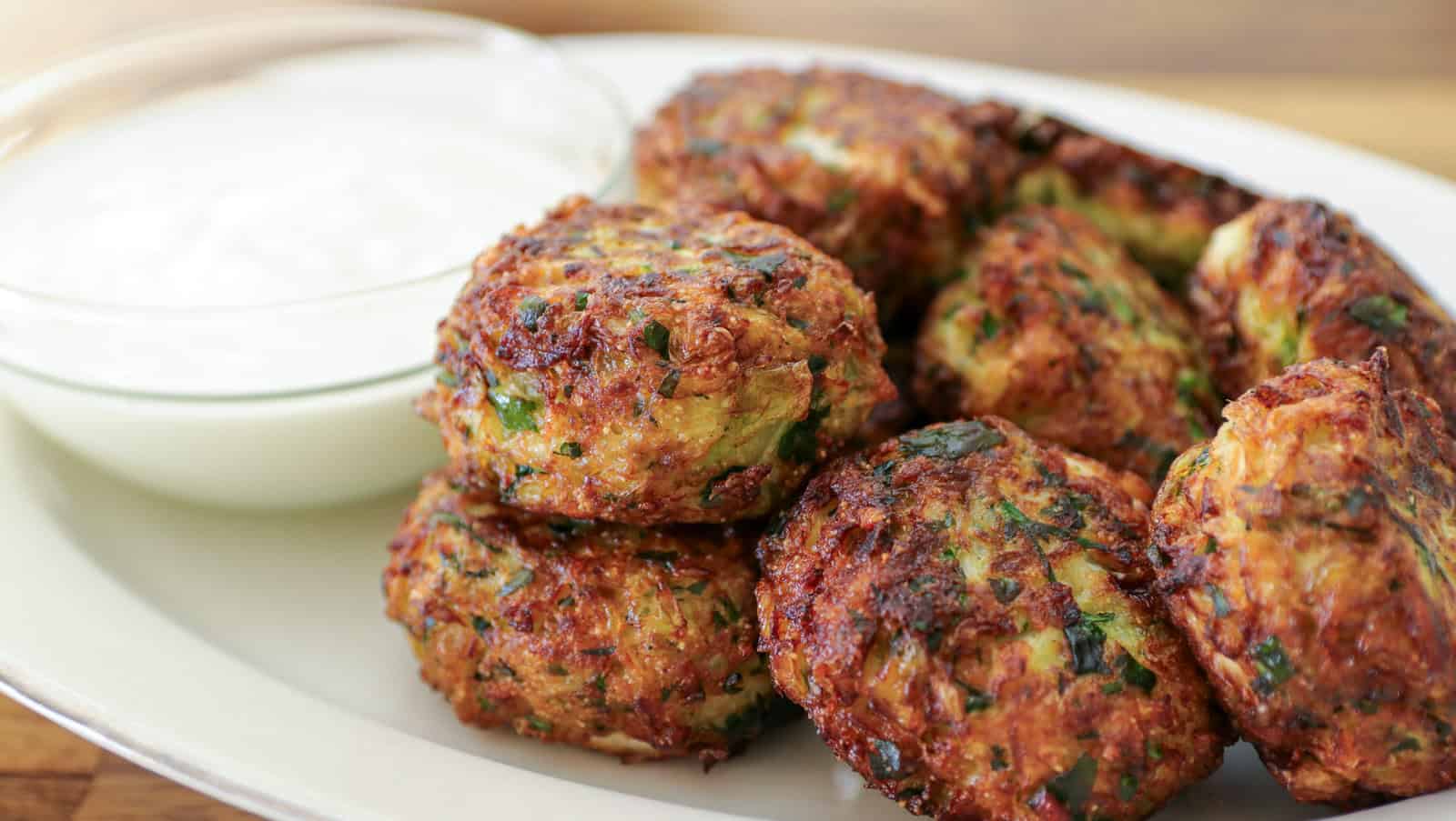 A plate of golden-brown zucchini fritters is stacked next to a small glass bowl filled with white yogurt sauce. The fritters are speckled with green herbs and have a crispy texture.