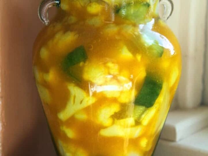 A jar of pickled vegetables in a yellow liquid. The jar contains a mix of cauliflower florets and cucumber pieces. It has a metal lid and is placed on a countertop against a light-colored wall and window frame.