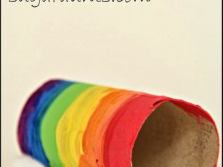 A rainbow craft made from a painted paper roll with vibrant stripes. The roll is adorned with cotton at both ends, resembling clouds. Text reads: Rainbow Craft sugaraunts.com.