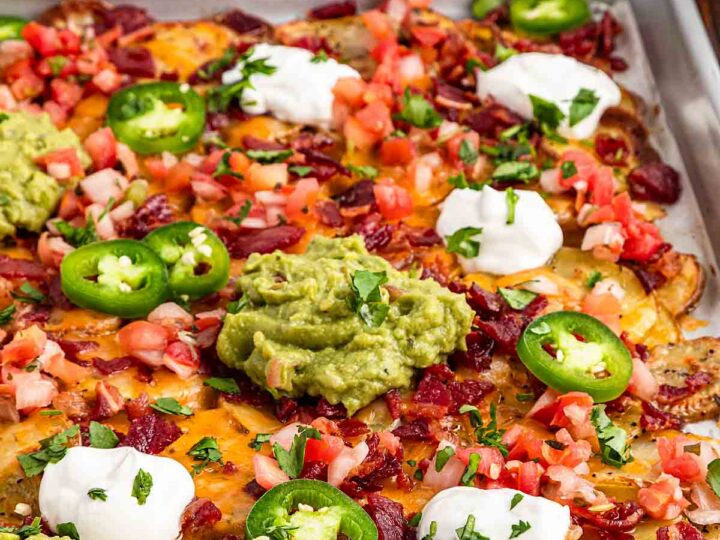 A tray of loaded nachos topped with melted cheese, sliced jalapeños, guacamole, sour cream, diced tomatoes, and chopped cilantro. Crispy bacon bits are scattered throughout, adding texture and flavor to the colorful dish.