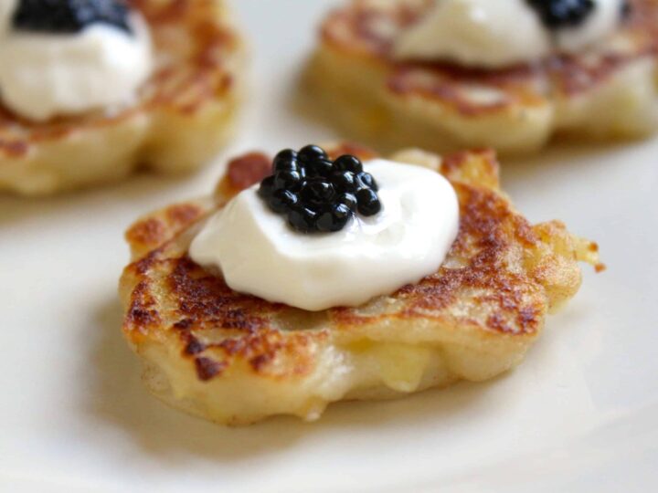 Close-up of a blini topped with a dollop of sour cream and black caviar on a white plate. The blini is golden brown and appears crispy. Other similarly garnished blinis are slightly blurred in the background.