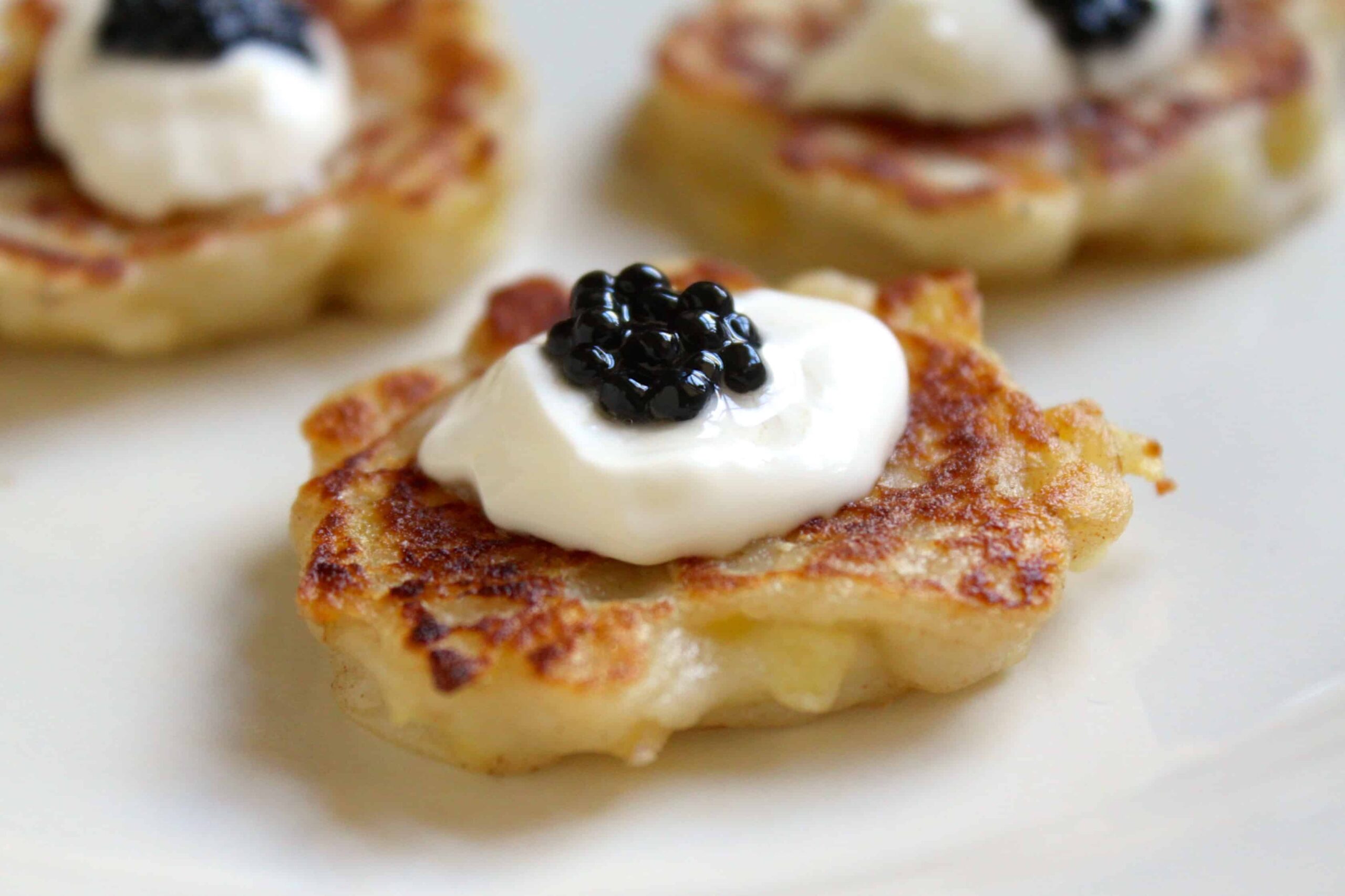 Close-up of a blini topped with a dollop of sour cream and black caviar on a white plate. The blini is golden brown and appears crispy. Other similarly garnished blinis are slightly blurred in the background.