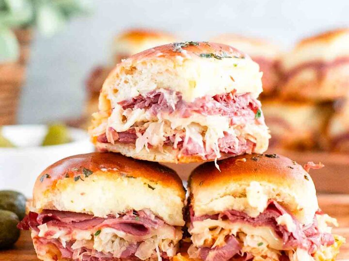 Three savory sliders stacked, featuring layers of corned beef, sauerkraut, and melted cheese between soft, golden buns. The sliders are garnished with herbs. A blurred background displays more sliders and a small dish with pickles.