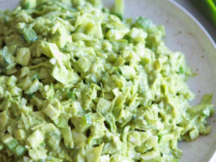 A plate of creamy avocado salad with finely chopped green vegetables, including cucumber and spring onions. Fresh green onions are visible in the background on a white surface.